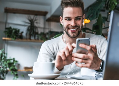 Portrait of smiling man with earphones sitting at coffee shop and using mobile phone. Young businessman holding having a business meeting conversation video call in a cafe. Freelance working concept