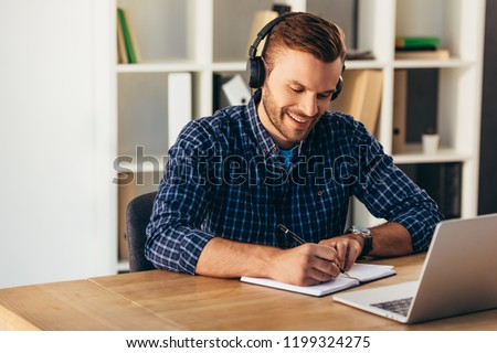 portrait of smiling man in headphones making notes while taking part in webinar at tabletop with laptop in office