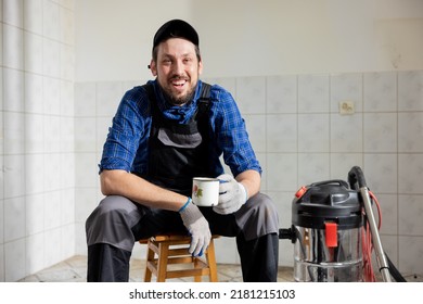 Portrait Of A Smiling Man Hired To Work On A Construction Site. The Man Is Sitting In A Room Undergoing Demolition Resting Drinking Coffee, Tea In A Break From The Renovations.