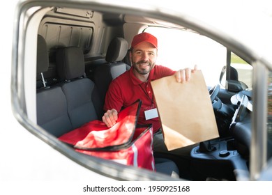 Portrait Of A Smiling Man In His 30s Delivering A Brown Bag With Lunch Or Dinner. Delivery Man Driving A White Van And Making A Home Delivery Service Of Takeout Food