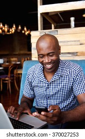 Portrait Of Smiling Man Looking At Mobile Phone While Sitting At A Cafe With A Laptop