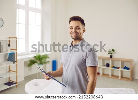 Portrait of smiling man physiotherapist with patient card in own cabinet in hospital. Happy male masseur or therapist ready for clients in wellness clinic. Healthcare and medicine concept.