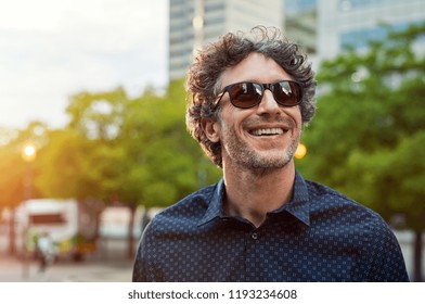 Portrait of smiling man wearing sunglasses and looking away in the city streets. Cheerful mature businessman walking with a big smile on face. Happy man in blue shirt and glasses enjoying the sunset. - Powered by Shutterstock