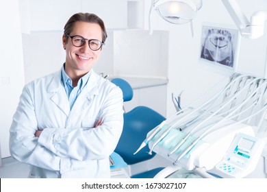Portrait of smiling man in white doctor robe uniform is standing in office, clinic with chair, instruments. Dentist workplace. Orthodontist is preparing for visit of patient. Dentistry concept.