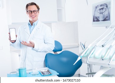 Portrait of smiling man in white doctor robe uniform, glasses is holding smartphone with blank screen, inviting patient to visit, standing in office, clinic. Dentist workplace. Dentistry concept. - Powered by Shutterstock