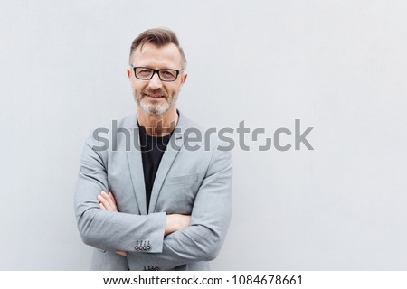 Portrait of smiling mature bearded man wearing glasses standing with arms crossed against bright background