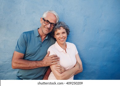 Portrait Of Smiling Mature Couple Standing Together Against Blue Background. Happy Middle Aged Man And Woman Against A Wall.