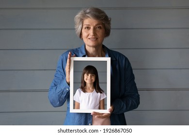 Portrait Of Smiling Mature Hispanic Grandmother Isolated On Grey Wall Background Show Granddaughter Picture In Frame. Elderly Senior Woman Hold Photo Of Her Photo As Child. Now And Then Concept.