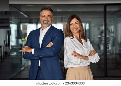 Portrait of smiling mature Latin or Indian business man and European business woman standing arms crossed in office. Two diverse colleagues, group team of confident professional business people. - Shutterstock ID 2343004303