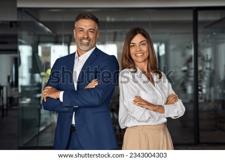 Portrait of smiling mature Latin or Indian business man and European business woman standing arms crossed in office. Two diverse colleagues, group team of confident professional business people.