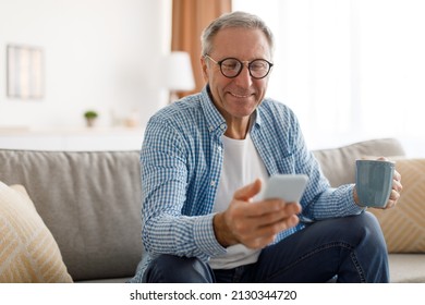 Portrait of smiling mature man in eyeglasses using mobile phone, watching video and holding cup of coffee, sitting on the couch in living room. Guy browsing internet, surfing web, free copy space - Powered by Shutterstock
