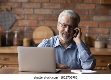 Portrait of smiling mature man sit at table at home talk on cellphone work on computer gadget. Smart senior 70s male have smartphone call conversation, use laptop. Elderly and technology concept. - Shutterstock ID 1822705982