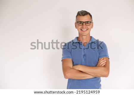 Portrait of smiling mature man standing with folded arms. Happy Caucasian man wearing blue T-shirt and eyeglasses standing over white background. Eyesight concept