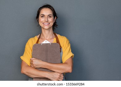 Portrait of smiling mature woman in apron holding clipboard and looking at camera. Beautiful waitress isolated on grey wall with copy space. Successful small business owner standing on grey background