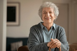 Portrait Of Smiling Mature Woman Holding Walking Stick And Sitting On Chair At Home. Portrait Of Happy Senior Woman Under Quarantine During Covid-19 Pandemic Smiling While Looking At Camera.