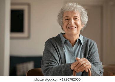 Portrait of smiling mature woman holding walking stick and sitting on chair at home. Portrait of happy senior woman under quarantine during covid-19 pandemic smiling while looking at camera.