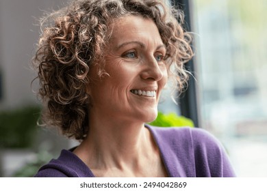 Portrait of smiling mature woman looking forwards near the window in the living room at home - Powered by Shutterstock