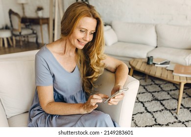 Portrait Of Smiling Mature Woman Lounging On White Sofa In Living-room At Home, Holding Phone Messaging Her Friends, Browsing Social Network Page, Receiving Funny Videos, Spending Leisure Time