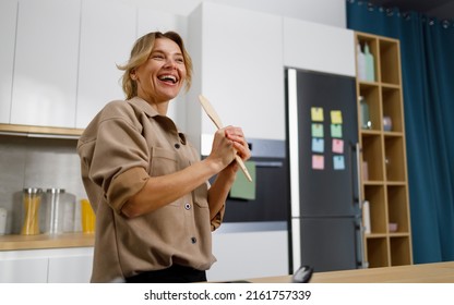 Portrait Of Smiling Mature Woman Singing In Kitchen With Wooden Spatula