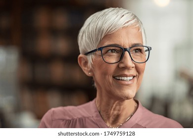 Portrait of smiling mature woman with spectacles in library looking away. Senior librarian standing in reading hall and thinking. Old beautiful lecturer contemplating. Future and vision concept.