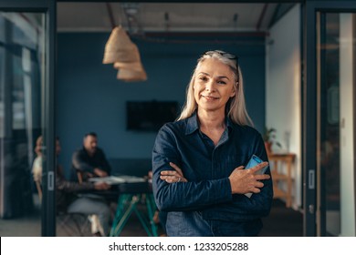 Portrait Of Smiling Mature Woman Standing Outside Meeting Room With Team Discussing Work In Background. Smiling Senior Business Woman At Office.