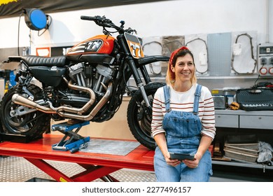 Portrait of smiling mechanic woman holding digital tablet sitting over platform with custom motorcycle on factory - Powered by Shutterstock