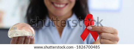 Portrait of smiling medical worker holding red ribbon cancer symbol on one hand. Silicone breast implant on other hand. Confidence and beauty. Modern medicine and womans body concept