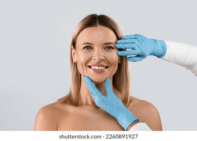 Portrait of smiling middle aged beautiful blonde woman getting prepared for plastic surgery procedures by doctor touching her face skin, isolated on grey background, copy space - Powered by Shutterstock