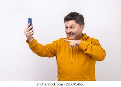 Portrait Of Smiling Middle Aged Man Pointing To Cell Phone Camera While Having Video Call Or Livestream, Wearing Urban Style Hoodie. Indoor Studio Shot Isolated On White Background.