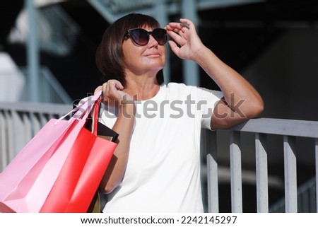 Portrait of smiling middle aged woman wearing stylish sunglasses and classic white t shirt holding colorful bags in hands after going stores. Fashion and shopping concept. Black Friday season sales.