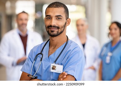 Portrait of smiling middle eastern man nurse with stethoscope looking at camera. Young doctor smiling while standing in hospital corridor with health care team in background. Successful indian surgeon - Powered by Shutterstock