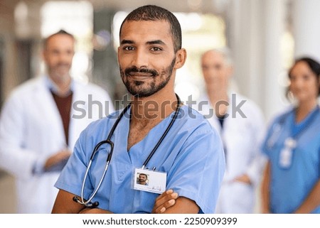 Portrait of smiling middle eastern man nurse with stethoscope looking at camera. Young doctor smiling while standing in hospital corridor with health care team in background. Successful indian surgeon