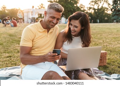 Portrait of smiling middle-aged couple man and woman holding cellphone while using laptop computer during picnic in summer park - Powered by Shutterstock