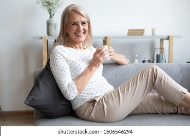Portrait of smiling middle-aged female retiree sit relax on cozy couch at home enjoy hot beverage, happy mature woman grandmother rest on comfortable sofa drink coffee or tea, look at camera posing