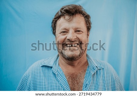 Portrait of a smiling middle-aged man with missing teeth and a beard, wearing a checkered shirt against a blue background.