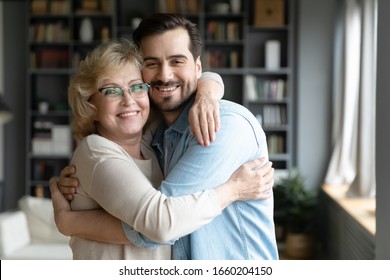Portrait of smiling middle-aged mother hug cuddle adult son relax in living room together, happy senior 70s mom embrace grown-up man child enjoy family weekend reunion at home, bonding concept
