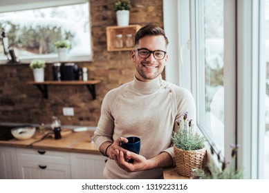 Portrait Of Smiling Millenial Man Drinking Tea Near The Window At Cozy Home On Winter Morning.