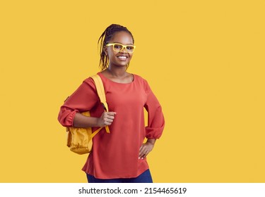 Portrait Of Smiling Millennial African American Girl Student In Glasses With Backpack Isolated On Yellow Studio Background. Happy Young Black Female University Or College Graduate. Education Concept.