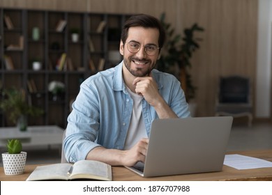 Portrait Of Smiling Millennial Caucasian Male Employee In Glasses Sit At Desk At Home Office Working Distant On Laptop. Happy Young 20s Man In Spectacles Use Computer, Consult Client Customer Online.