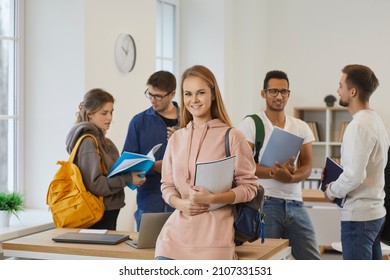 Portrait Of Smiling Millennial Teen Girl Student With Backpack And Book In University With Mates. Happy Young Caucasian Teenager Learner In School Or College With Groupmates. Education Concept.