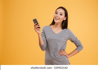 Portrait Of Smiling Model With Phone And Arm At Hip. Isolated Orange Background