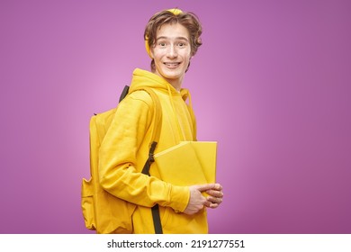 Portrait Of A Smiling Modern Teenage Boy In Bright Youth Clothes Standing With Books And A Backpack On A Purple Background. Youth Fashion. Educational Concept. Copy Space.