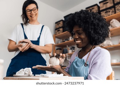 Portrait of smiling multicultural female pottery class student sitting at pottery workshop with clay in hands and learning clay work with instructor. Diverse pottery course attendee learning pottery. - Powered by Shutterstock