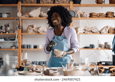 Portrait of smiling multicultural pottery designer standing at ceramics studio with earthenware in hands and laughing. Cheerful interracial female potter having fun making earthenware at workshop. - Powered by Shutterstock