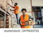 Portrait of smiling multicultural storage worker with box in hands smiling at camera at facility and his colleague in background working.