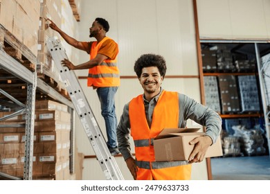 Portrait of smiling multicultural storage worker with box in hands smiling at camera at facility and his colleague in background working. - Powered by Shutterstock