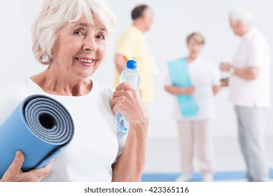 Portrait Of A Smiling Old Lady Holding An Exercise Mat And A Bottle Of Water, With Her Friends In The Blurred Background