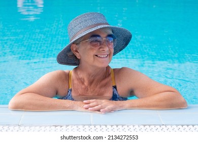 Portrait Of Smiling Old Senior Woman Leaning On The Edge Of Swimming Pool. Caucasian Carefree Lady Wearing Blue Hat And Sunglasses Enjoying Free Time And Vacation In The Pool Water