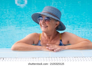 Portrait Of Smiling Old Senior Woman Leaning On The Edge Of Swimming Pool. Caucasian Carefree Lady Wearing Blue Straw Hat And Sunglasses Enjoying Free Time And Vacation In The Pool Water