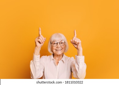 Portrait Of Smiling Old Woman With Short Gray Hair And Round Glasses Wearing White Blouse, Pointing Fingers Up. Senior Woman Isolated Over Orange Background